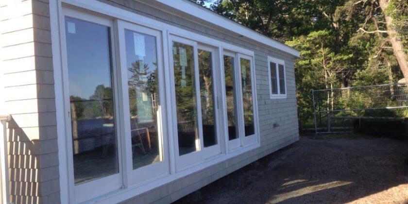 A sunroom addition designed by a Boston contractor, with multiple windows on a residential home, overlooking a yard with trees in the background.