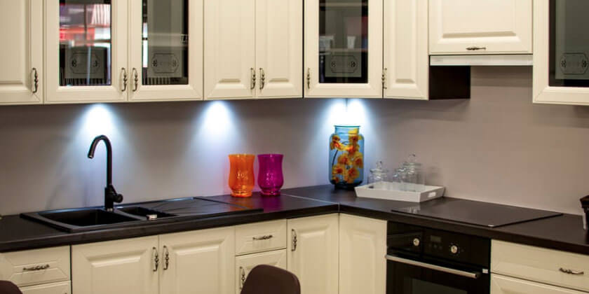 Modern kitchen with under-cabinet lighting, featuring a black sink, white cabinets, and colorful glassware on the countertop, designed by a renowned Boston contractor.