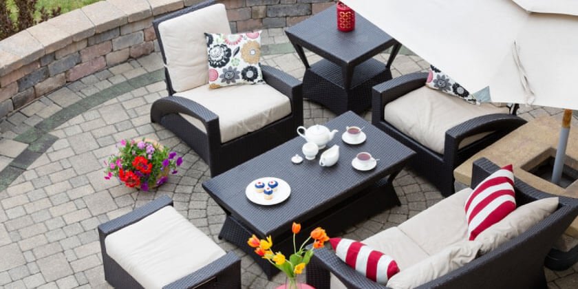 Patio area with wicker furniture set, including a sofa, chairs, and a table with a tea set, decorated with colorful cushions, under an open umbrella, surrounded by pavers and a p